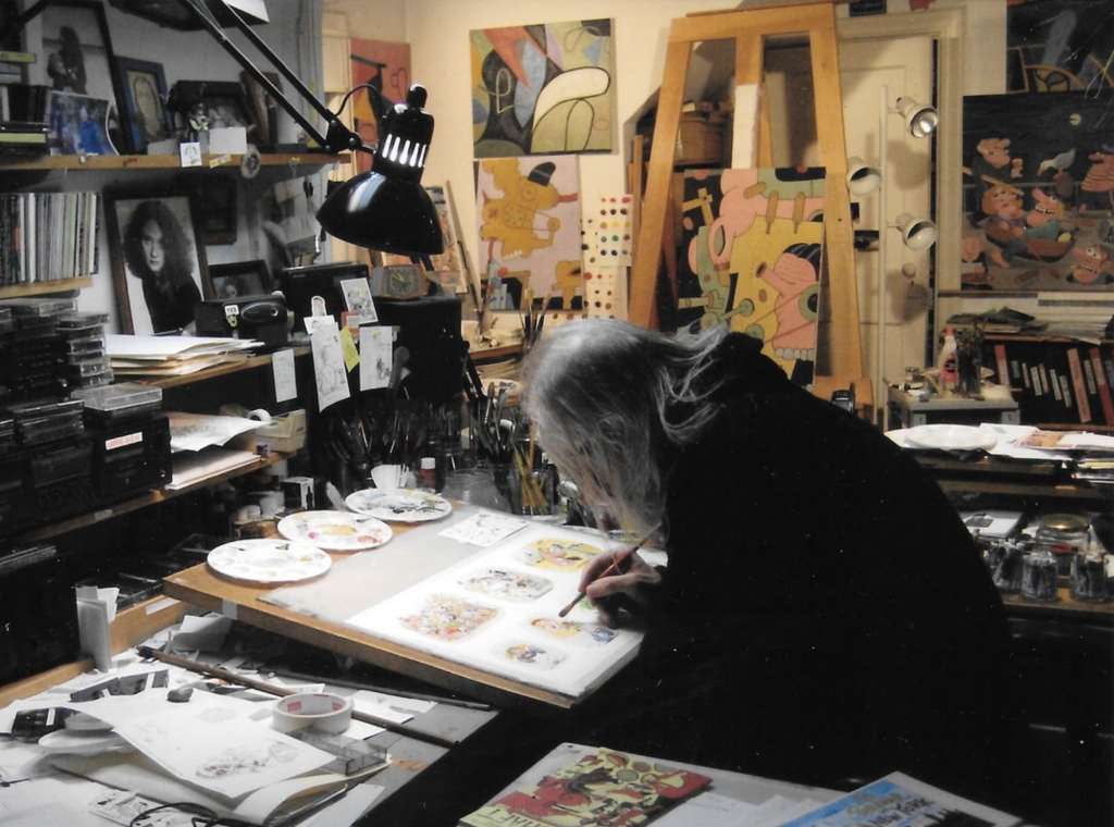 Photographie de Tom Bunk dans son atelier new-yorkais, penché sur une planche d’illustrations à son bureau. L’espace est saturé de peintures, d’objets, de palettes et de livres, reflétant un univers foisonnant, chaotique et minutieux à l’image de son œuvre.