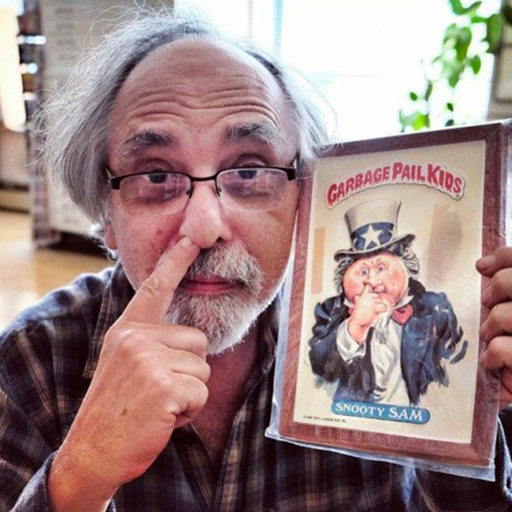 Art Spiegelman en 2015 posant avec une carte Garbage Pail Kids, le doigt dans le nez en clin d’œil à l’illustration qu’il tient.