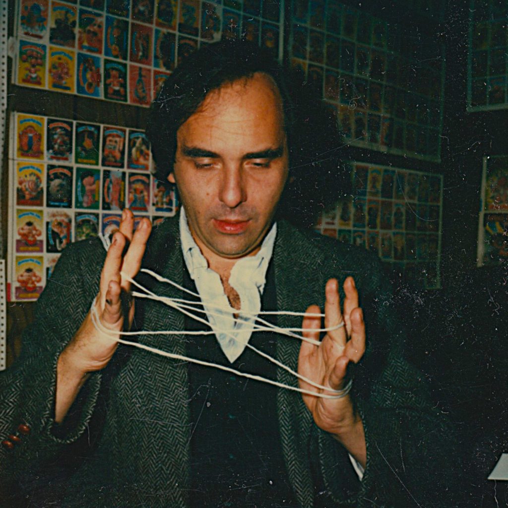 Art Spiegelman dans les bureaux de Topps en 1987, concentré sur un jeu de ficelles, en arrière-plan une collection de cartes Garbage Pail Kids tapisse le mur.