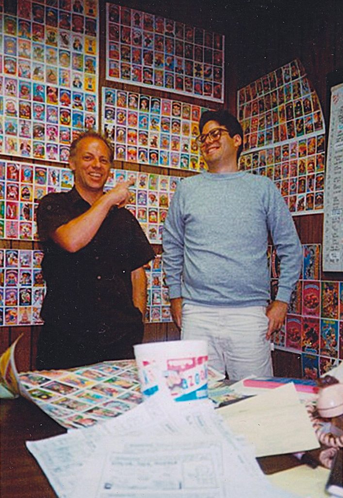 Tom Bunk (à gauche) et Mark Newgarden (à droite) posent devant un mur tapissé de cartes Garbage Pail Kids chez Topps, dans une ambiance joyeuse des années 80.