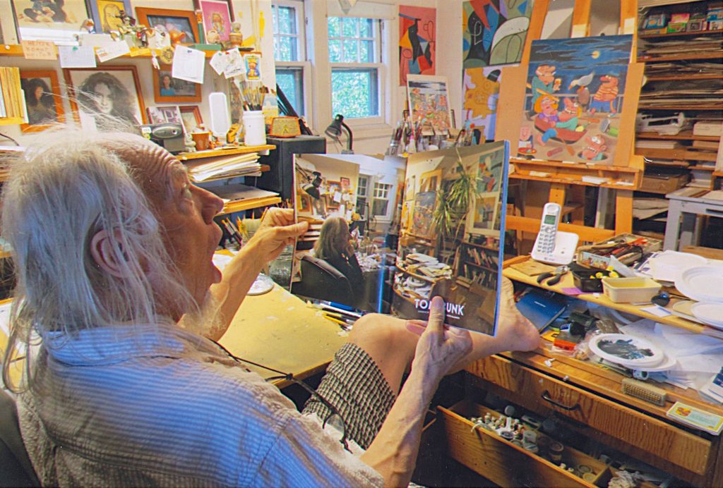Tom Bunk dans son atelier à Brooklyn, lisant un livre ouvert à une double page le représentant lui-même dans ce même atelier, entouré de ses peintures, pinceaux et outils de création.