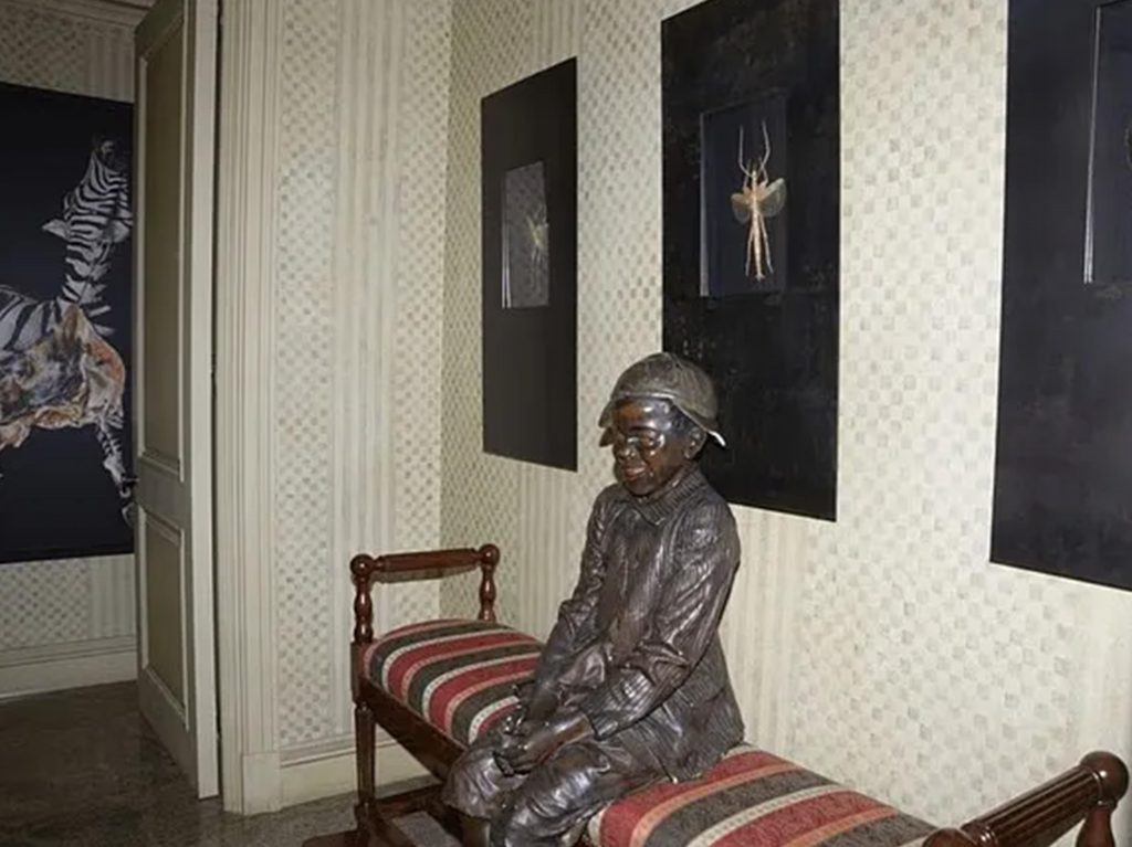 Statue en bronze représentant un enfant assis sur un banc dans un couloir décoré de tableaux encadrés et de papier peint clair.