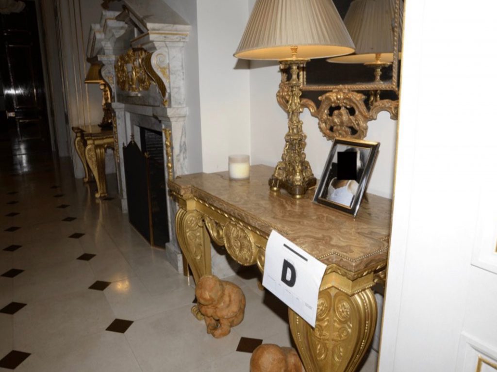 Console en marbre et dorures dans un couloir, avec lampe, cadre photo et deux petites statuettes d’enfants posées au sol devant le meuble.