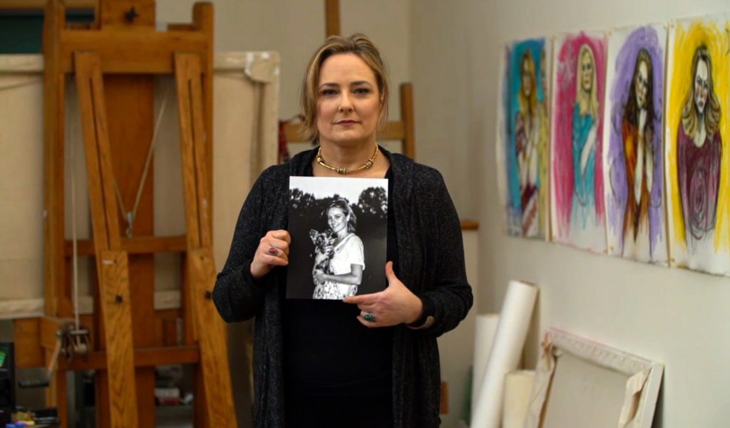 Maria Farmer debout dans son atelier d’artiste, tenant une photographie en noir et blanc d’elle plus jeune ; derrière elle, plusieurs portraits colorés de femmes accrochés au mur, issus de sa série “The Survivors Project”