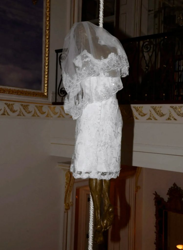 Gros plan d’une sculpture suspendue : figure féminine portant une robe blanche et un voile, accrochée à une corde à l’intérieur d’un escalier résidentiel.