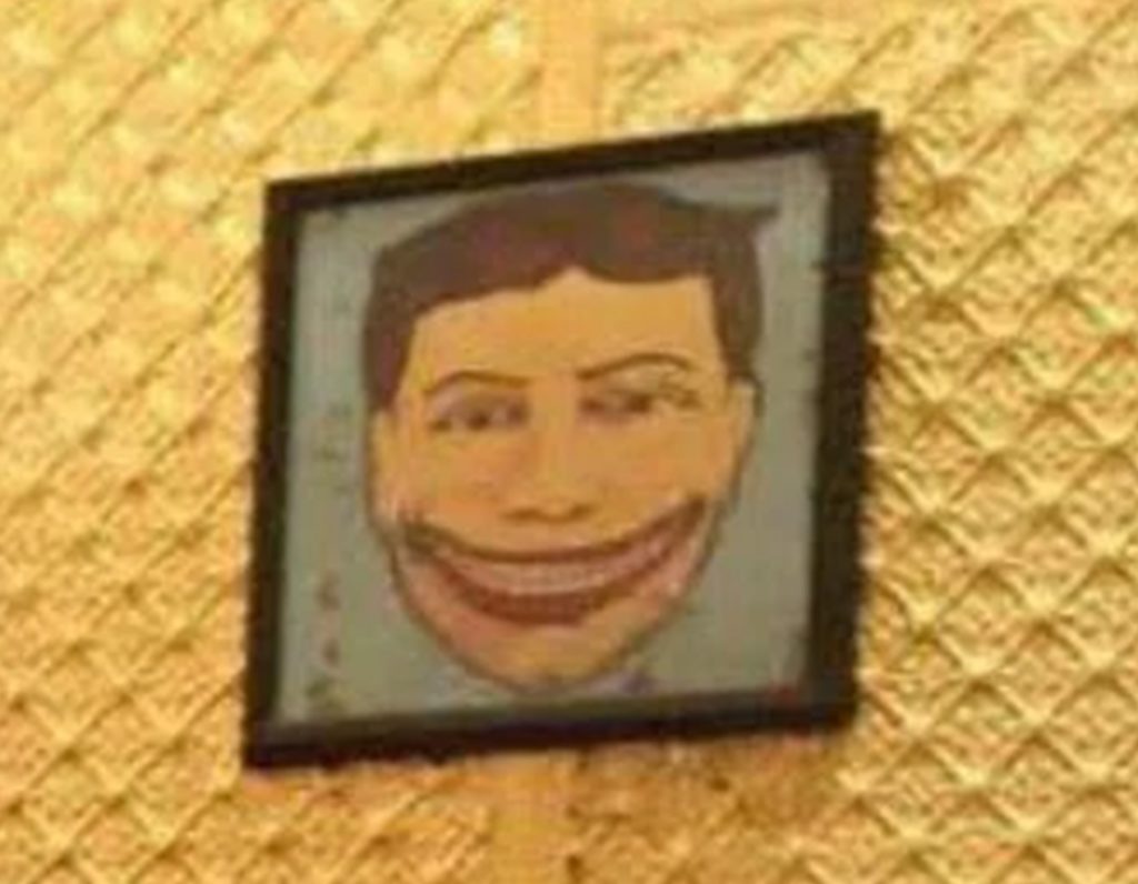 Photographie d’un tableau encadré représentant le Steeplechase Face, un visage masculin stylisé au large sourire exagéré, accroché sur un mur à motif doré.