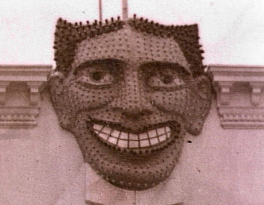 Photographie ancienne en sépia d’un grand visage sculpté accroché à une façade, avec yeux fixes et large sourire aux dents apparentes, emblème du Steeplechase Park à Coney Island.