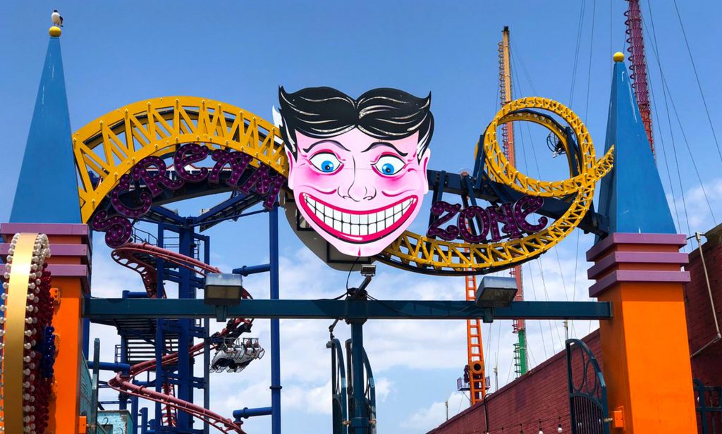 Enseigne moderne “Scream Zone” à Coney Island avec un grand visage stylisé aux yeux écarquillés et au large sourire blanc, entouré de rails de montagnes russes jaunes et rouges sous un ciel bleu.