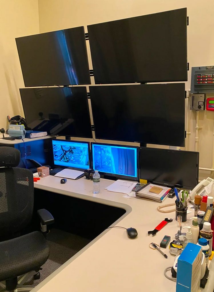 Bureau équipé d’un mur d’écrans noirs, avec plusieurs moniteurs superposés au-dessus d’un plan de travail, évoquant un espace de vidéosurveillance.