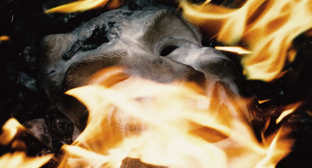 Masque blanc de théâtre en train de brûler dans les flammes, sur fond sombre, pour la série photo Mascarade.