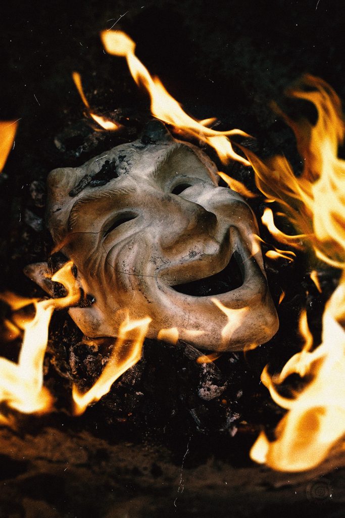 Photographie des faux-semblants : masque blanc de théâtre en train de brûler sur fond sombre.